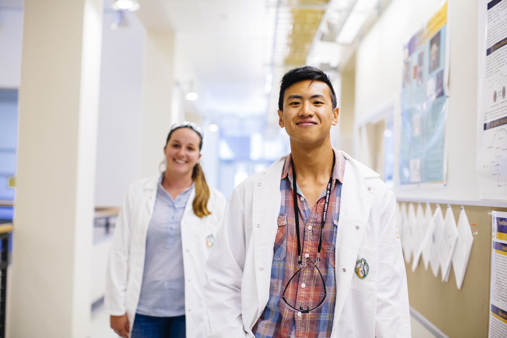 Two student in white lab coats walking down a hallway smiling.